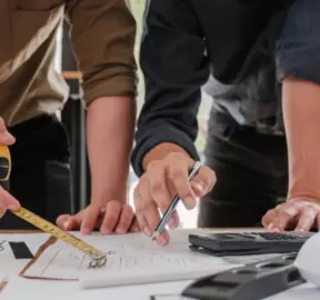 Due persone intente a prendere le misure e a discutere i progetti di costruzione su una scrivania, utilizzando un metro a nastro, una penna e una calcolatrice. Sono visibili solo le loro mani e le loro braccia, con l’attenzione concentrata sullo spazio di lavoro collaborativo: una scena che mette in risalto l’importanza della formazione nel settore edile, poiché un lavoro di squadra preciso è essenziale per il successo. Due persone intente a prendere le misure e a discutere i progetti di costruzione su una scrivania, utilizzando un metro a nastro, una penna e una calcolatrice. Sono visibili solo le loro mani e le loro braccia, con l’attenzione concentrata sullo spazio di lavoro collaborativo: una scena che mette in risalto l’importanza della formazione nel settore edile, poiché un lavoro di squadra preciso è essenziale per il successo.