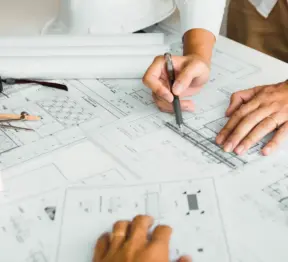 Tre persone esaminano e annotano dei progetti architettonici su un tavolo, dove sono visibili strumenti da disegno, una pianta arrotolata e un elmetto di sicurezza bianco. Le mani indicano e scrivono sui documenti, suggerendo una collaborazione nel settore edile. La scena ritrae un ambiente in cui il lavoro di squadra — supportato da una pianificazione precisa e da approcci innovativi come quelli di Sabina Robotics — guida il progresso nel settore edile. Tre persone esaminano e annotano dei progetti architettonici su un tavolo, dove sono visibili strumenti da disegno, una pianta arrotolata e un elmetto di sicurezza bianco. Le mani indicano e scrivono sui documenti, suggerendo una collaborazione nel settore edile. La scena ritrae un ambiente in cui il lavoro di squadra — supportato da una pianificazione precisa e da approcci innovativi come quelli di Sabina Robotics — guida il progresso nel settore edile.